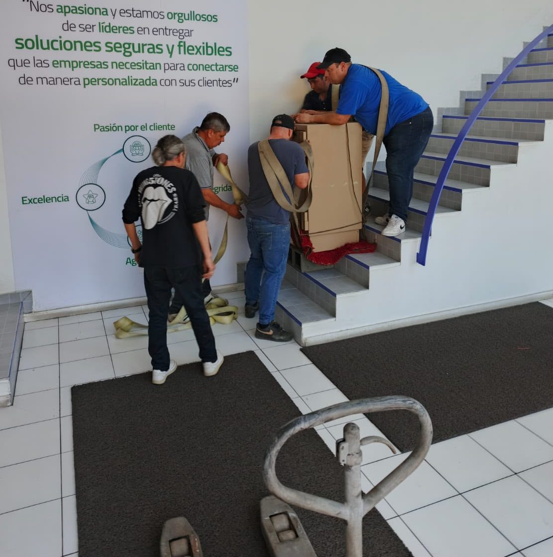 Cinco técnicos con eslingas bajan una caja fuerte embalada en cartón por la escalera de una empresa