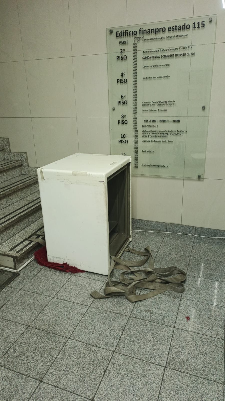 Caja fuerte blanca en el hall de un edificio de oficinas tras bajar la escalera, con eslingas en el piso