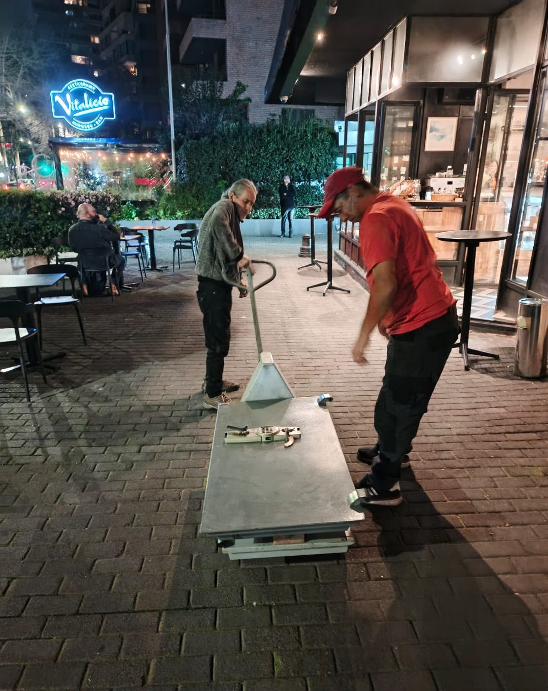 Dos técnicos maniobran una caja fuerte acostada sobre transpaleta en la vereda, de noche